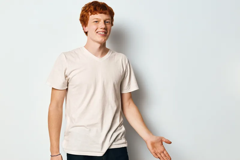 Young man with red hair wearing cream t shirt standing and smiling with open hand gesture on white background in studio. Casual expression and relaxed body language.