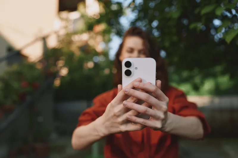 Young woman in a red shirt takes a selfie outdoors, holding a smartphone close to her face as the sun lights her relaxed expression with a soft breeze ruffling hair and a blurred background