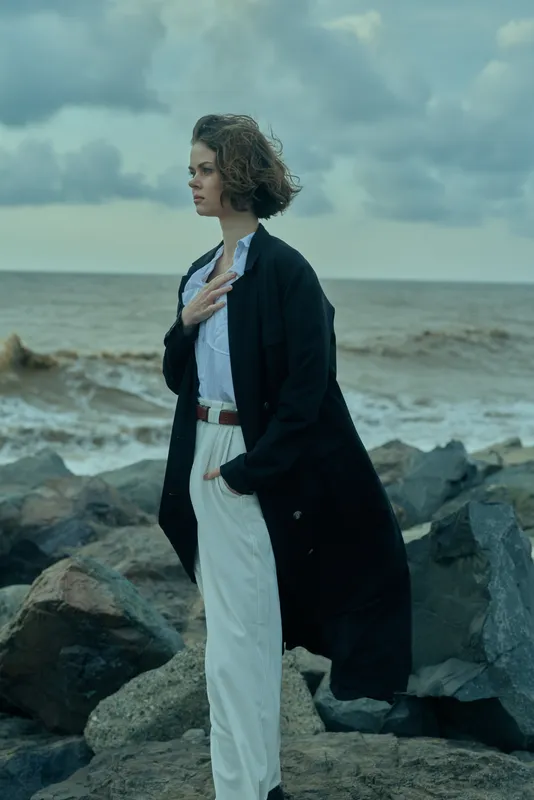 Windblown coat, sea breeze and rocky shoreline set a dramatic scene as a stylish woman gazes into the distance, hand on chest, calm mood, monochrome palette, coastal fashion