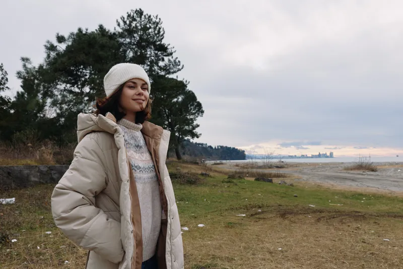 Winter woman outdoors wearing a thick coat and knit hat stands near a lakeside shore, calm sky, trees in distance, casual fashion style for outdoor weather photography.