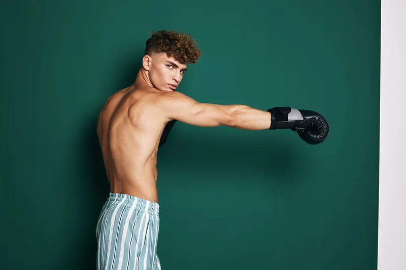Young man with curly hair wearing boxing gloves and striped pants training with punch against green wall in studio. Athletic shirtless male boxer practicing martial arts and fitness.