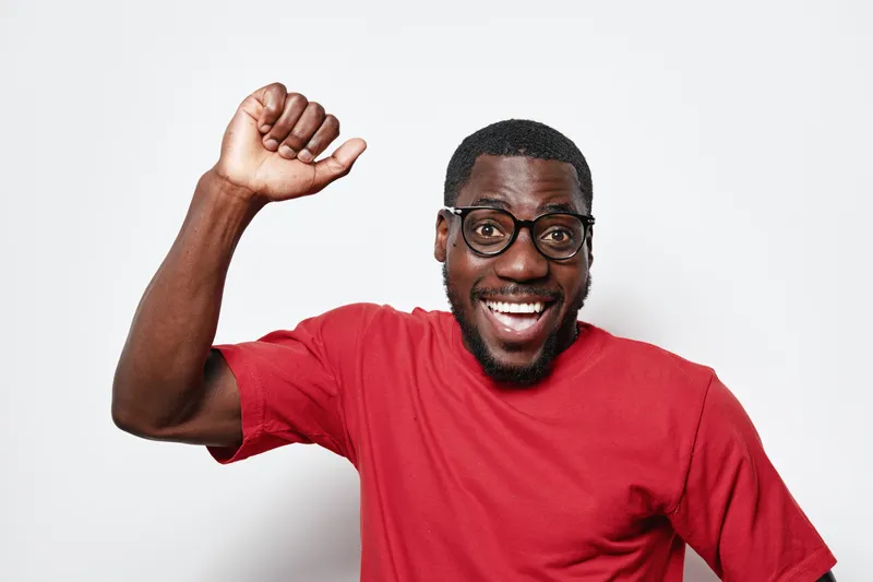 African American man with dark skin wearing glasses and red t shirt smiling and raising fist in celebration or success on white background. Positive emotion and casual style.