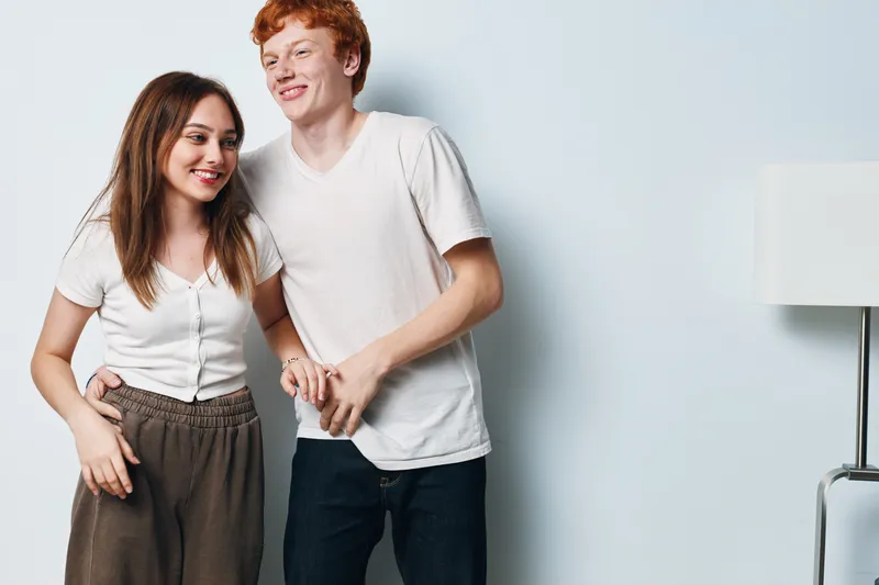 Young man and woman smiling and holding hands standing near white wall indoors. Casual clothing with happy mood and natural light in lifestyle portrait of friendship and connection.