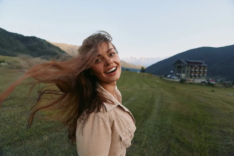 Woman lifestyle outdoor in meadow with mountains, wind in hair, beige jacket, smiling, enjoying nature, bright natural light, carefree mood, scenic rural setting, soft warm glowing colors in the