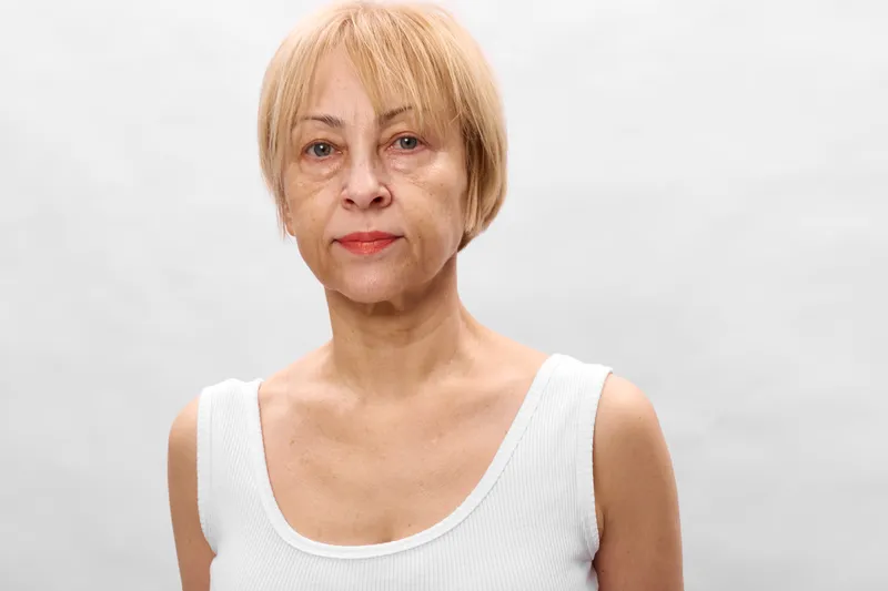 Senior woman with short blonde hair in a studio headshot facing camera. Calm expression with red lipstick and natural makeup on light gray background. Beauty and aging concept.