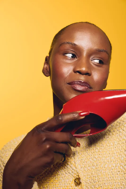 Close-up of a thoughtful black woman with dark skin holding a red high heel near her face, wearing a yellow sweater. Concept of fashion, beauty, and style on yellow background.