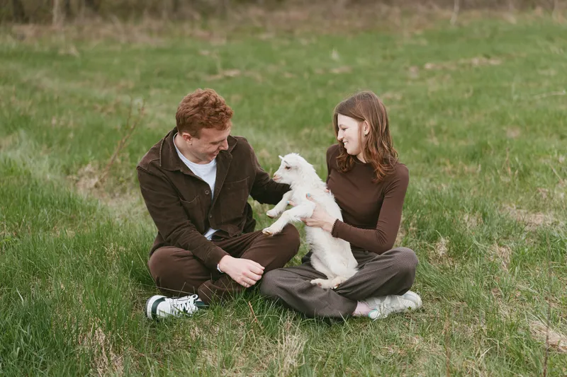 Smiling couple sitting on grass outdoors with a small goat kid, interacting gently. Warm daylight, casual clothing, relaxed mood, and natural setting conveying lifestyle, companionship, and animal