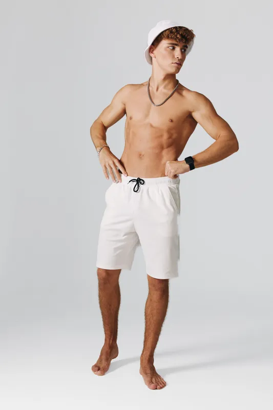 Young man standing barefoot in white shorts and bucket hat looking to side with hands on hips against plain background. Casual summer style with accessories and relaxed pose.