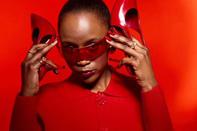Dark skinned African American woman with short hair holds red high heels near head wearing red sunglasses and red outfit on red background. Concept of fashion, style, and confidence.