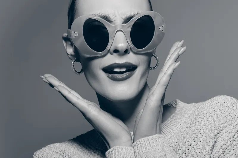 Happy white woman with light skin wearing large sunglasses and earrings poses with hands near face in studio. Concept of fashion and joyful expression.