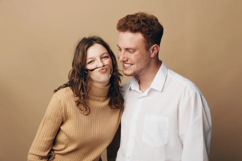 Young couple with playful mood, woman making mustache with hair, man smiling and looking at her. Casual clothing and warm beige background create cozy and intimate atmosphere.