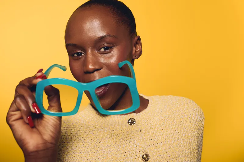 Confident black woman with dark skin holds turquoise eyeglasses close to camera on yellow background. Studio portrait of African female model wearing beige knit sweater with positive expression.