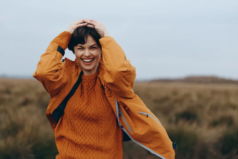 Woman smiling outdoors in autumn countryside, wearing orange coat, hands on head in a field, lifestyle moment with warm light, natural landscape, candid joyful portrait