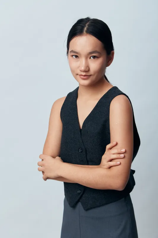 Young woman standing with arms crossed looking confident at camera wearing dark sleeveless vest and skirt with straight black hair on light gray background in studio.