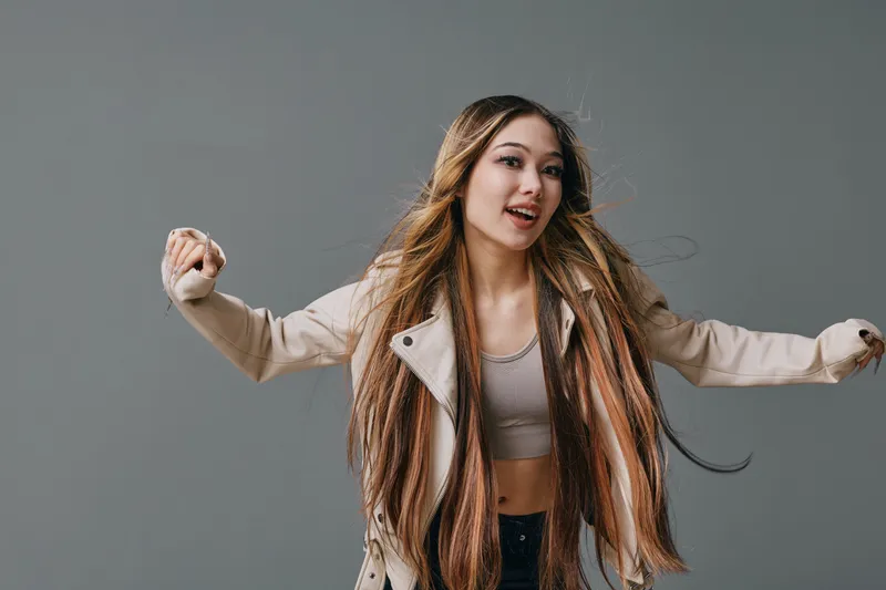 Young woman with long braided hair wearing beige jacket and crop top posing and dancing in studio with gray background, smiling and expressing joy and freedom.