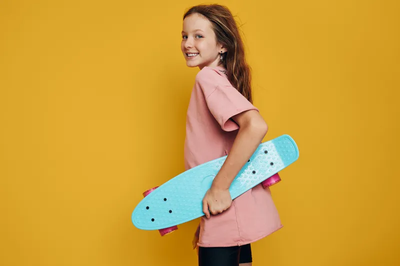Smiling girl holding a blue skateboard standing against a yellow background. Casual clothing and confident posture. Youth lifestyle concept.