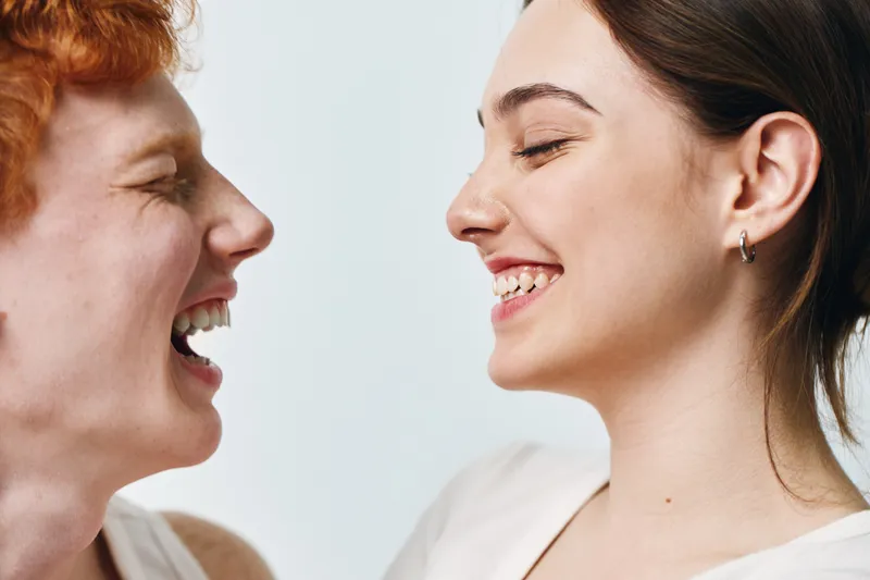 Two women smiling and looking at each other with happy expressions closeup portrait for friendship and positive emotions in studio with light background and natural skin tones