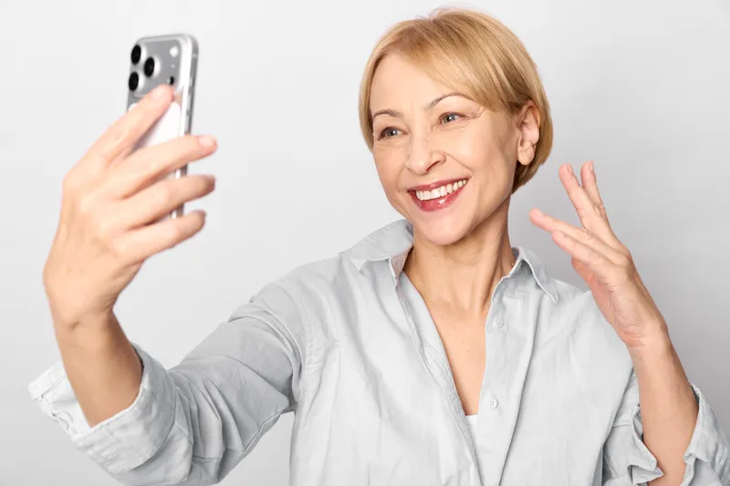 Selfie smiling senior woman holds phone, waving to camera, joyful portrait indoors, smartphone closeup, light minimal background, casual gray shirt smile wide