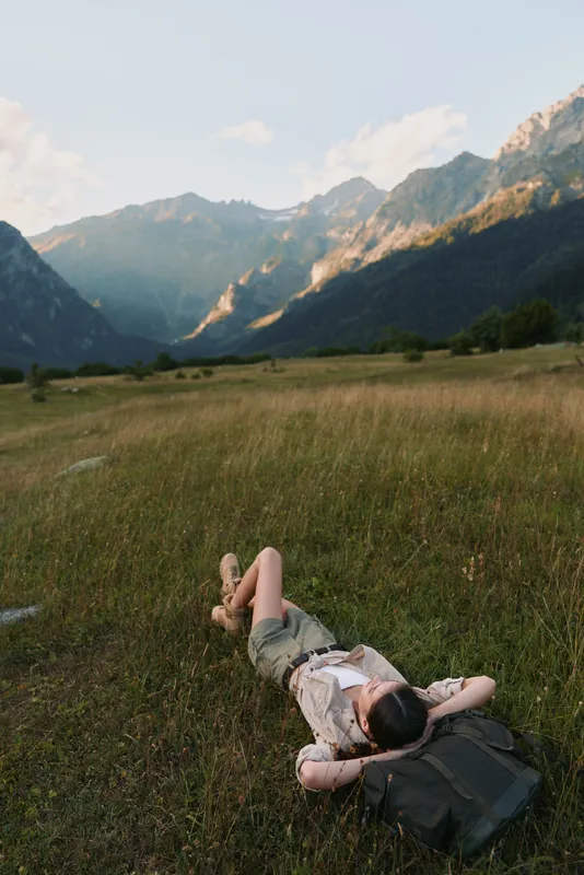 A woman lies on grass in a mountain valley with hands behind head, relaxing and enjoying nature. The scene shows peaceful lifestyle and outdoor leisure in a scenic landscape.