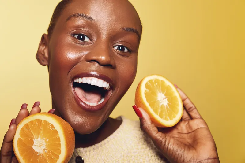 Happy African woman with dark skin holding two halves of an orange fruit, smiling widely against yellow background. Concept of healthy lifestyle, vitamin, and natural beauty.