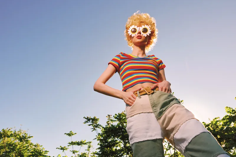 Woman wearing a striped top, patchwork pants, and floral glasses stands outdoors under bright sun. Retro film color mood, lifestyle vibe, casual mood, playful and confident.