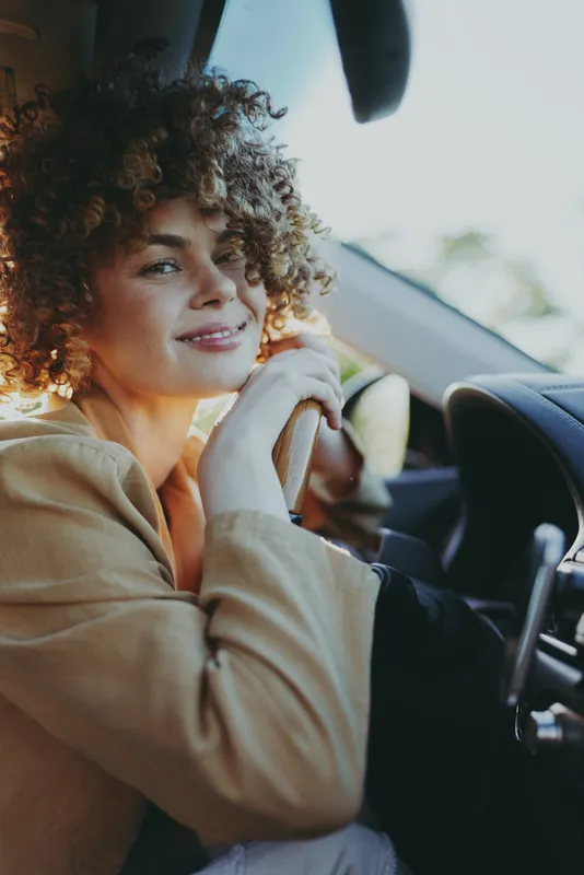Woman driving a car in a sunlit moment, relaxed posture, candid smile, interior details visible, lifestyle vibe with natural colors and soft breeze, capturing everyday freedom and warm mood outdoors