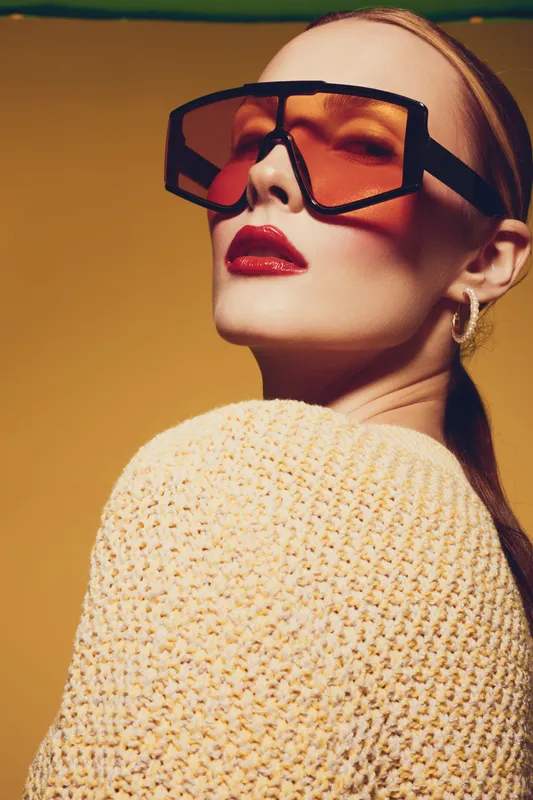 Stylish woman with oversized orange sunglasses and red lipstick poses confidently against yellow background. She wears a textured sweater and gold hoop earrings. Fashion and beauty concept.
