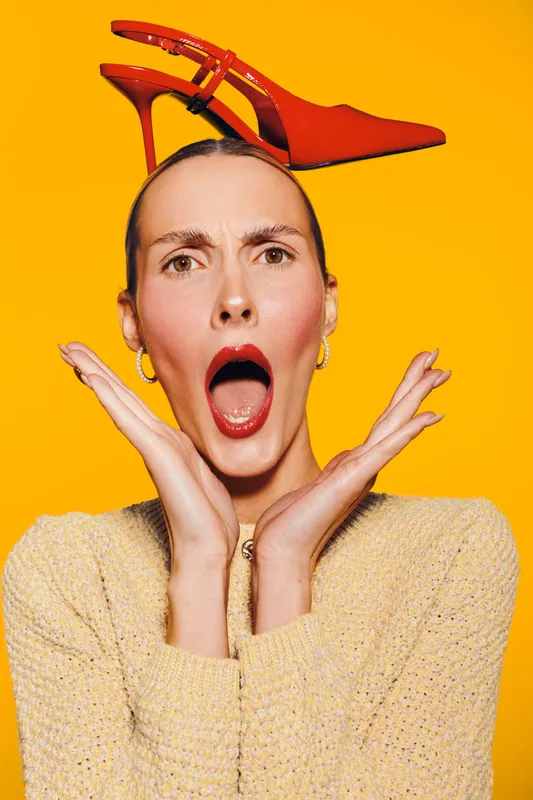 Surprised woman with red lipstick and earrings holds red high heel shoe on head against bright yellow background. Concept for fashion, style, and unexpected moments.