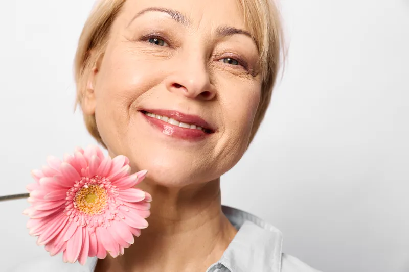 Smiling blonde woman holds a pink gerbera flower near cheek, soft light closeup portrait on white background, calm beauty and freshness, minimal studio look.