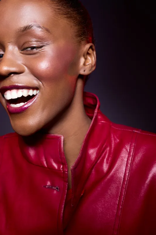 Close-up of happy African woman with dark skin laughing, wearing shiny red leather jacket. Studio portrait with dark background. Concept of joy, fashion, and confident beauty.