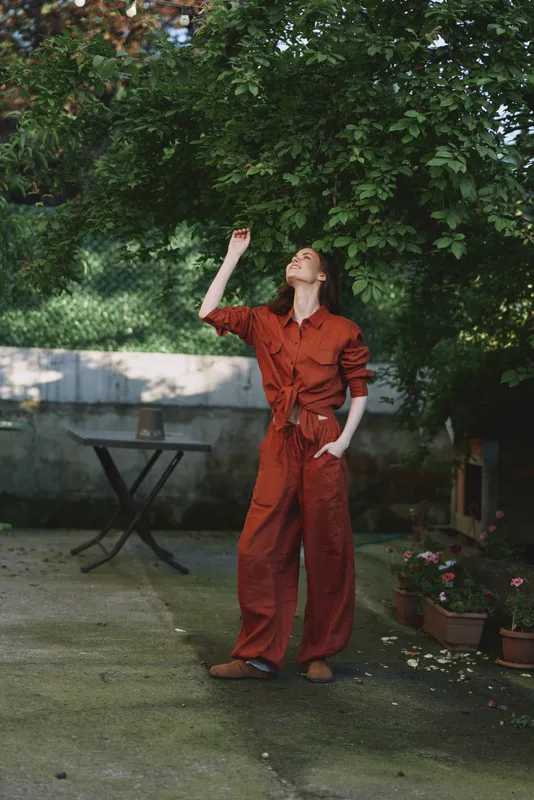 A woman in a red jumpsuit stands outdoors, looking up with a raised hand. A bright garden setting conveys casual daily life, playful fashion, and personal style