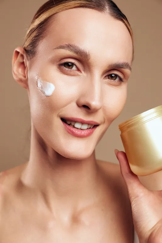Young woman applies face cream on cheek holds jar of moisturizer smiling with clean skin natural makeup isolated on beige background beauty skincare routine concept.