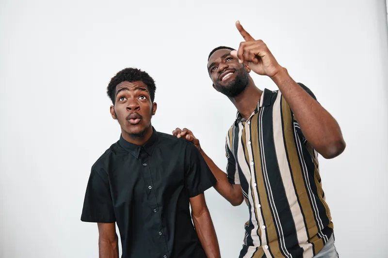 Two African American men with dark skin standing together indoors, one pointing up and the other looking surprised, casual clothing and friendly interaction on white background.