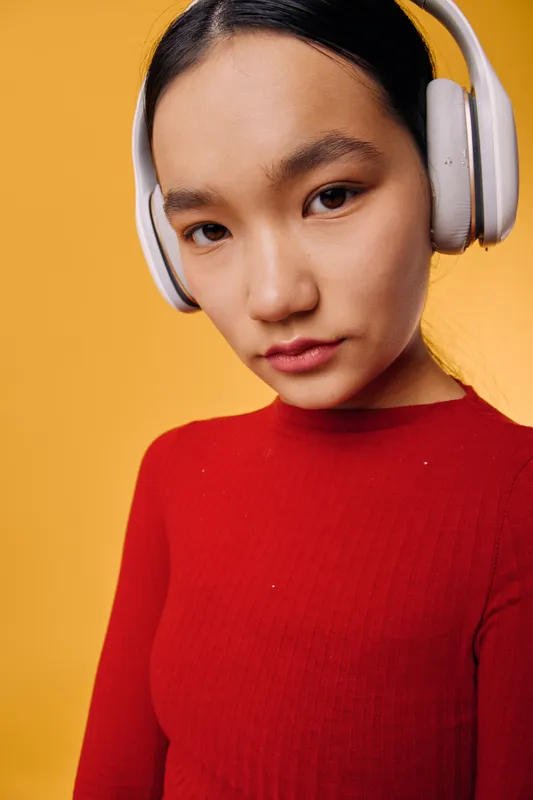 Woman wearing white headphones and red sweater, looking at camera with neutral expression on yellow background. Closeup portrait of female listening to music or podcast at studio indoors.