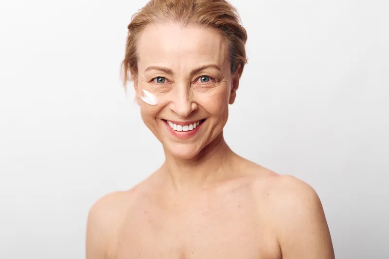 Smiling middle aged woman with natural skin applies white face cream on cheek, isolated on white background. Concept of skincare, aging beauty, self care, and healthy lifestyle.