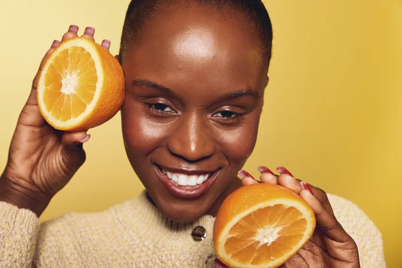 Happy African woman with dark skin holds two halves of fresh orange fruit near her face. Smiling female model in beige sweater on yellow background. Concept of health, freshness, and beauty.