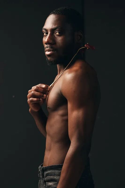 African man with dark skin and muscular body posing shirtless in creative conceptual style against dark background. Artistic portrait with flower on shoulder and confident expression.