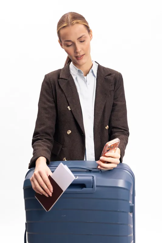 Young woman in brown jacket holds passport and smartphone while standing with blue suitcase. Concept of travel, vacation planning, and modern lifestyle with luggage on white background.