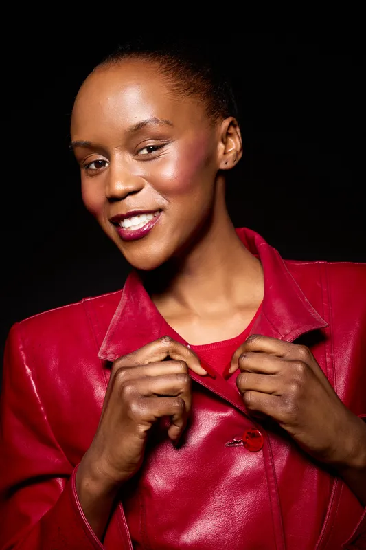 Confident African woman with dark skin wearing a red leather jacket smiles brightly against a black background. Concept of fashion, beauty, and empowerment in studio lighting.