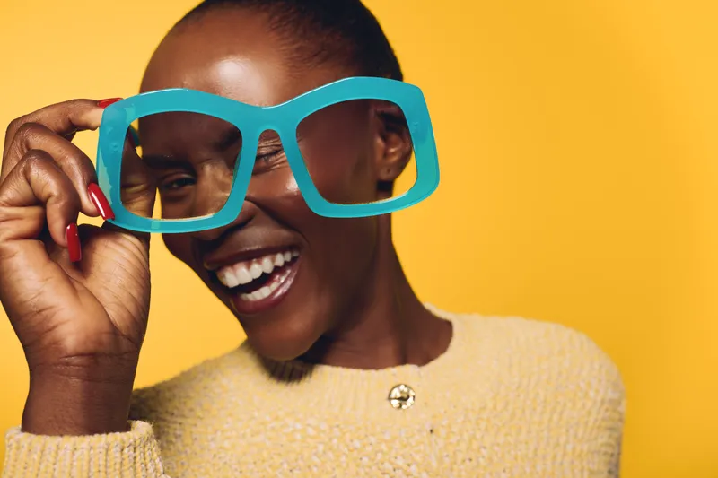 Happy dark skinned African woman smiling broadly holds oversized blue glasses near face. Yellow background and beige sweater create bright cheerful mood. Concept for joy and fashion.