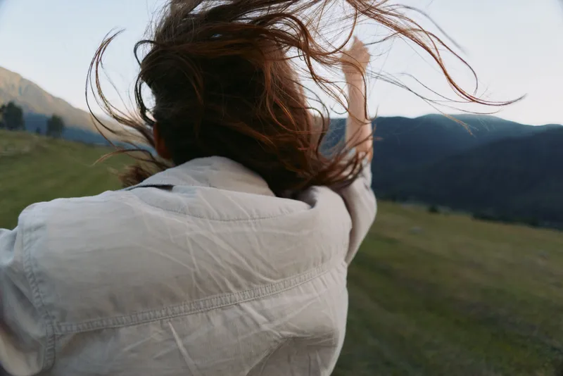 Young woman with long hair wearing a white shirt raises her arm outdoors in a mountainous landscape during daylight, expressing freedom and connection with nature.