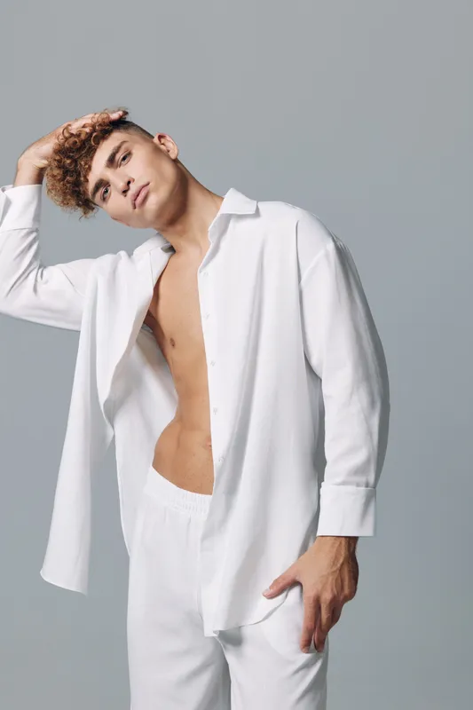 Young man with curly hair wearing white open shirt and pants posing with hand on head against gray background in studio. Fashion, casual style and confident expression.