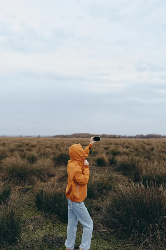 Woman outdoors in a field of grass, orange jacket, lifestyle smartphone moment, holding a smartphone, standing in nature, cloudy sky, calm relaxed posture
