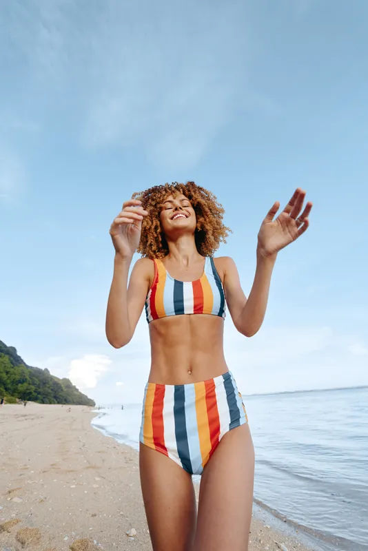 Happy woman with curly hair wearing colorful striped bikini standing on sandy beach near calm sea under clear sky, enjoying sunny day and fresh air outdoors.