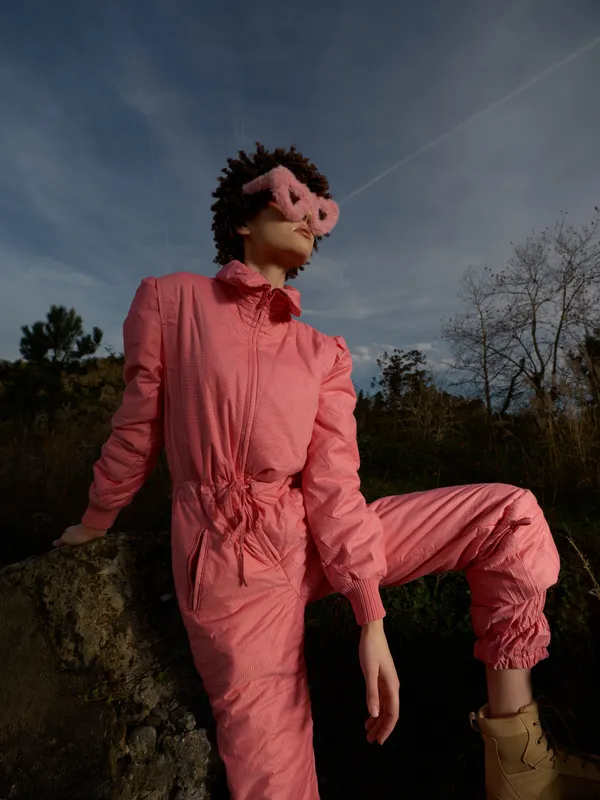 Pink jumpsuit and pig mask wearer posed outdoors on a rocky ledge under a blue sky, creating a bold fashion portrait with surreal texture and dramatic lighting.