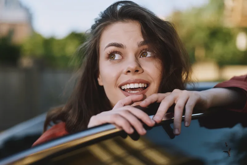 Smiling woman with long hair resting her hands on a car window, outdoor scene, bright sunlight, lifestyle vibe, casual confidence and warmth, candid moment that evokes everyday wellness happiness