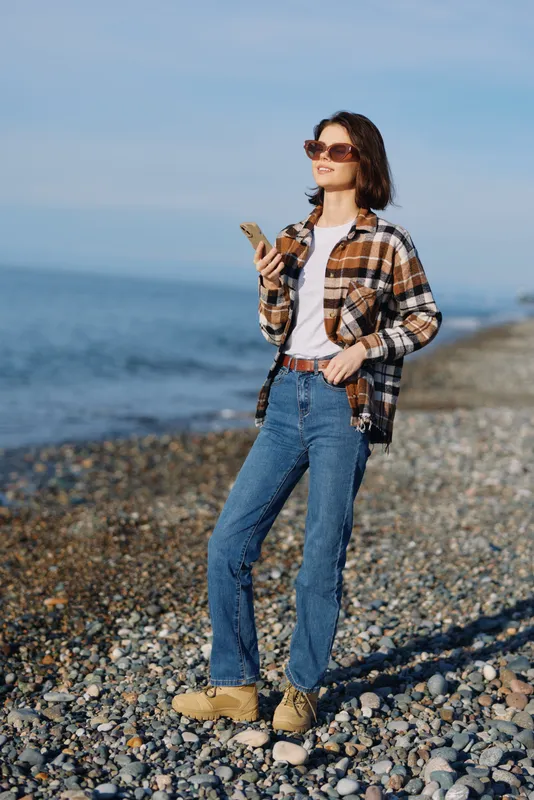 Woman on a pebble beach using a smartphone, dressed in casual plaid shirt, jeans and boots, enjoying seaside scenery with clear sky and calm water.
