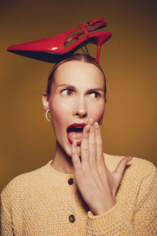 Surprised woman with red lipstick and hoop earrings wears a red high heel shoe on her head. She covers her mouth with her hand against a warm brown background. Concept of fashion and creativity.