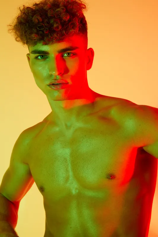Young man with curly hair and toned muscles posing shirtless in colorful studio lighting with red and green hues on skin, looking at camera, fashion and beauty concept.