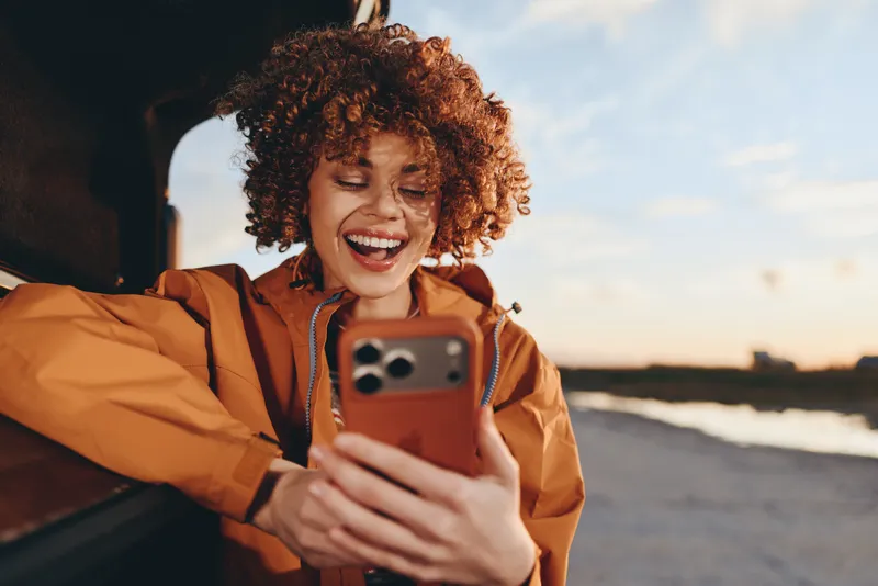 Woman lifestyle, curly hair, orange jacket, smiling while holding smartphone near beach at sunset, warm light, coastal sky, candid travel moment, carefree mood.